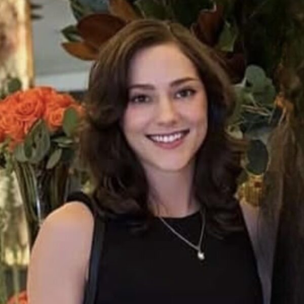 http://Headshot%20of%20Courtney%20Arkbar%20smiling%20in%20black%20dress%20with%20necklace,%20flowers%20and%20plants%20in%20background.