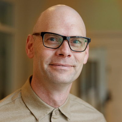 http://Headshot%20of%20Patrick%20Rashleigh%20from%20shoulders%20up,%20smiling%20in%20tan%20button%20down%20with%20glasses.