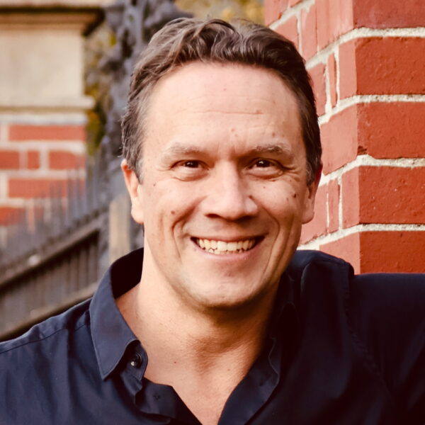 http://Headshot%20of%20Lin%20Fisher%20from%20shoulder%20up%20with%20arm%20raised,%20smiling%20in%20dark%20shirt,%20brick%20wall%20and%20fence%20in%20background.