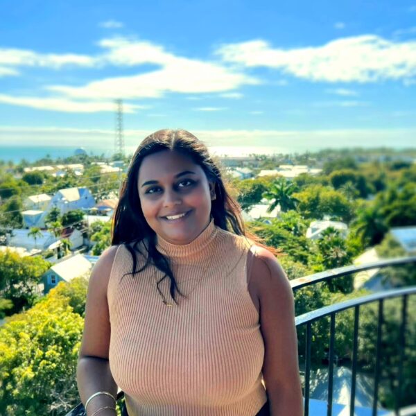 http://Tarika%20Sankar%20in%20tan%20top,%20smiling%20and%20looking%20at%20camera,%20standing%20on%20a%20balcony%20overlooking%20a%20sunny%20coastal%20town%20with%20palm%20trees%20and%20the%20ocean%20in%20the%20distance.